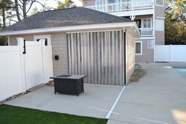 An outdoor patio featuring a black fire pit, a beige building with accordion metal shutters, and white privacy fencing.