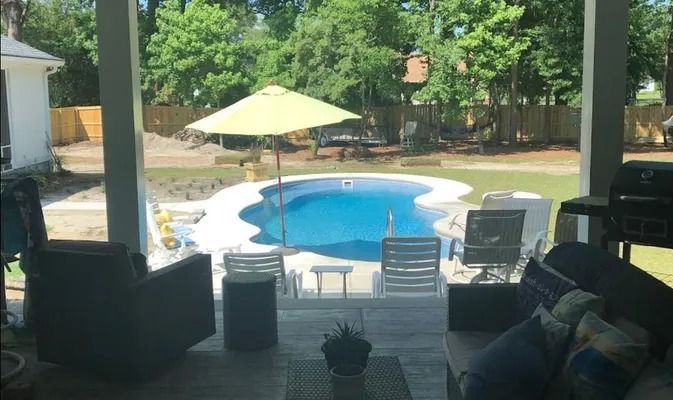 View from a shaded porch looking out toward a blue kidney-shaped swimming pool in a backyard with a yellow umbrella.