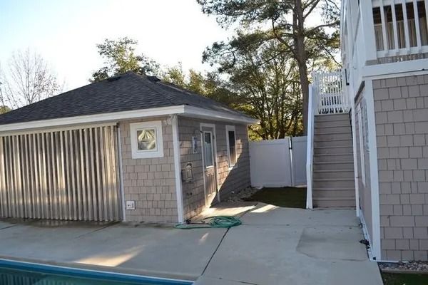 A beige shed with a grey shingled roof stands next to a pool deck and a staircase leading to a multi-story house.