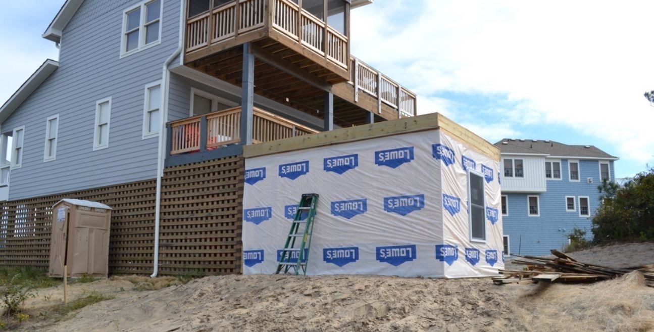 A home addition under construction with white house wrap, a ladder, and surrounding sandy terrain.
