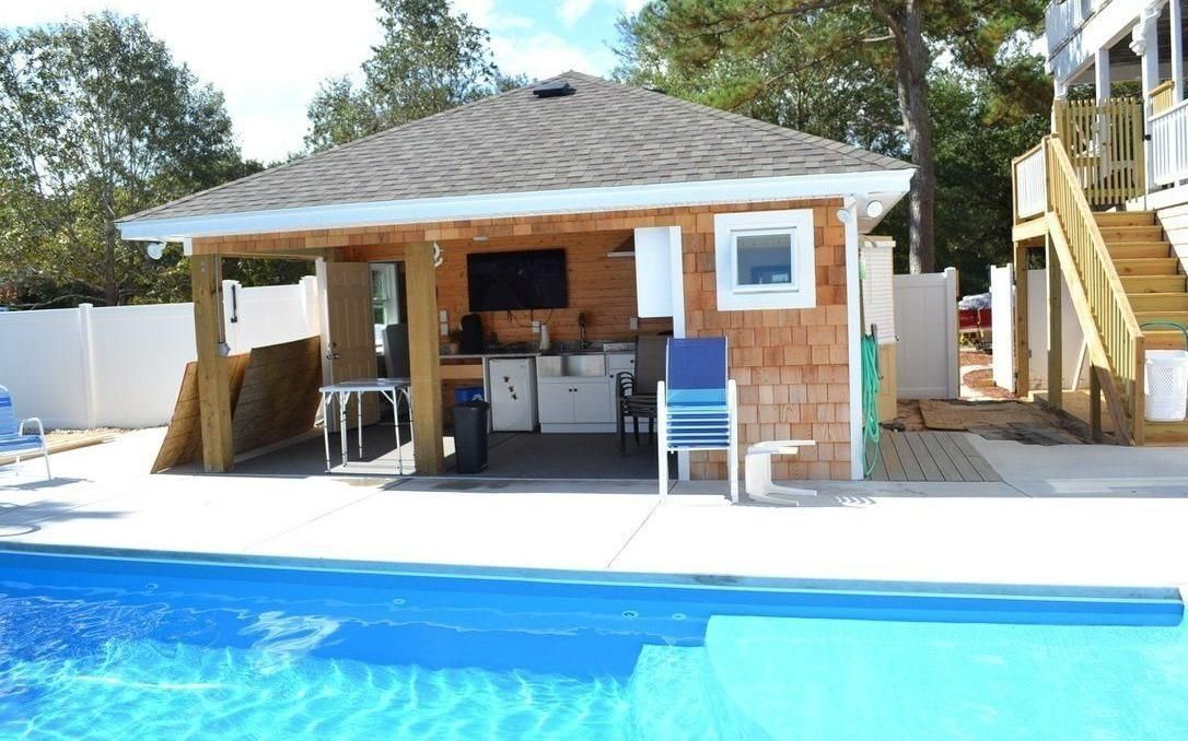 A poolside cedar-shingled cabana featuring a wet bar, outdoor television, and seating, set on a bright concrete patio.