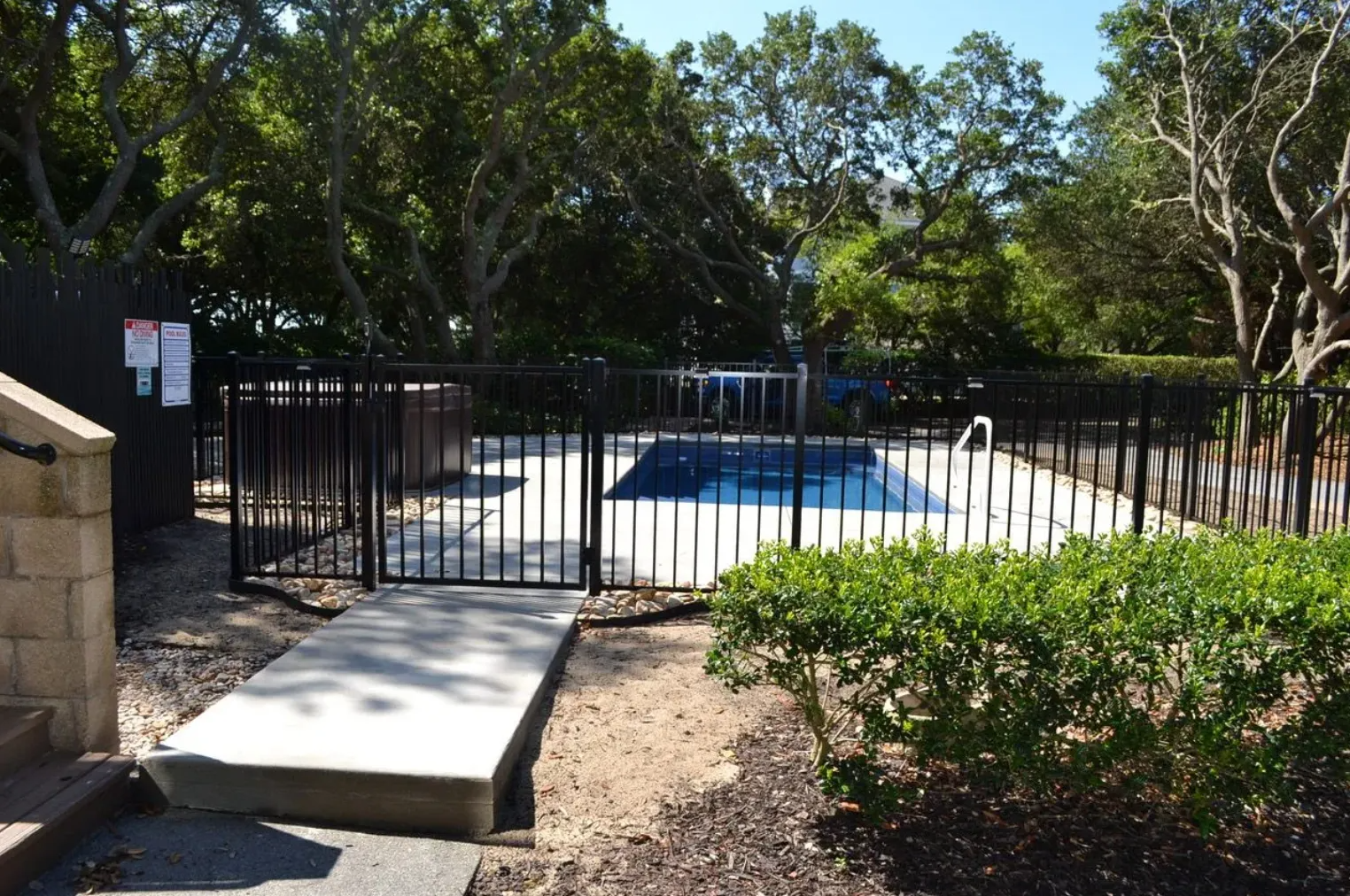 A black metal-fenced outdoor pool and hot tub area surrounded by green trees on a sunny day.