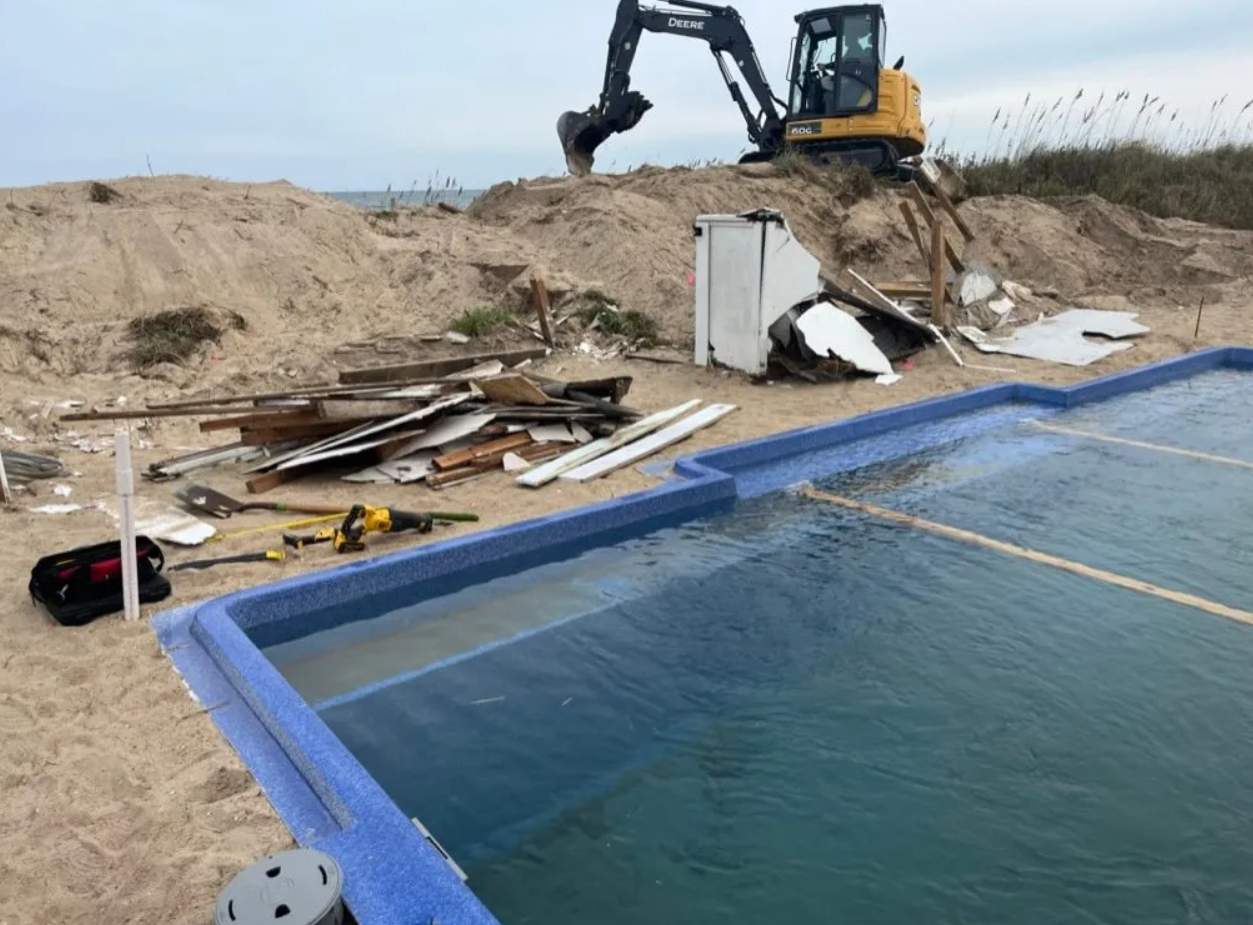 A yellow excavator works on a sandy beach near a swimming pool filled with debris and construction waste.
