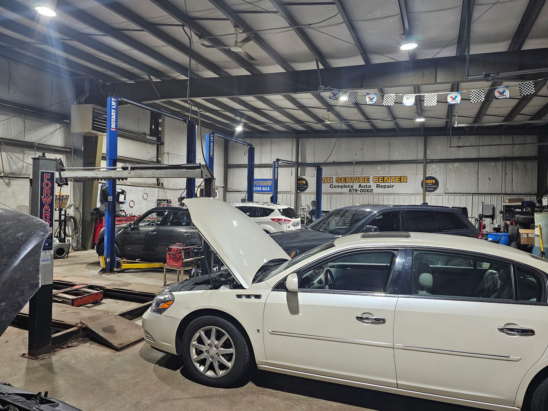 Auto repair shop with two cars on lifts, one white sedan with hood open in the foreground.