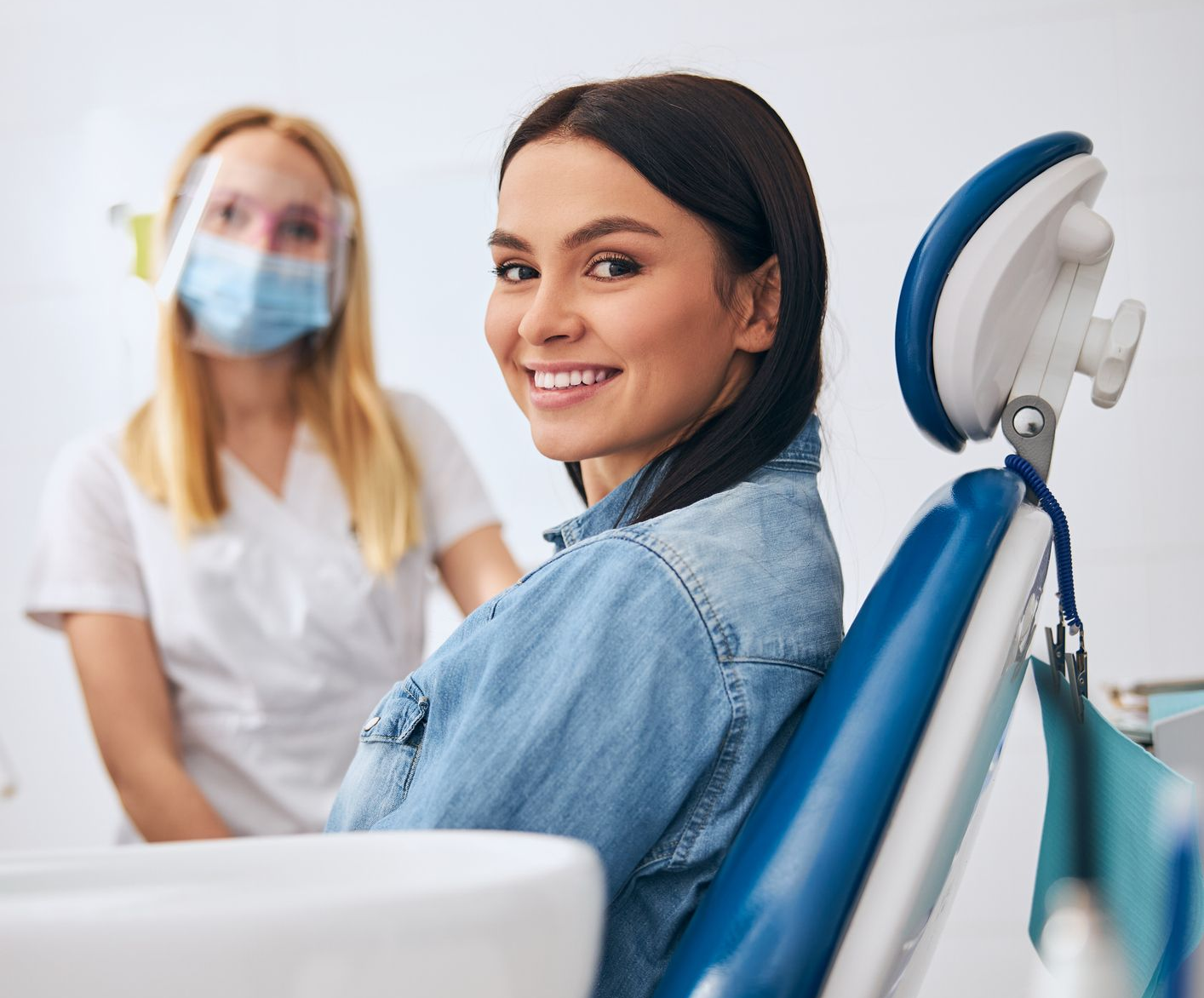 Smiling patient sitting in dental chair at modern dental centre with dentist in background.