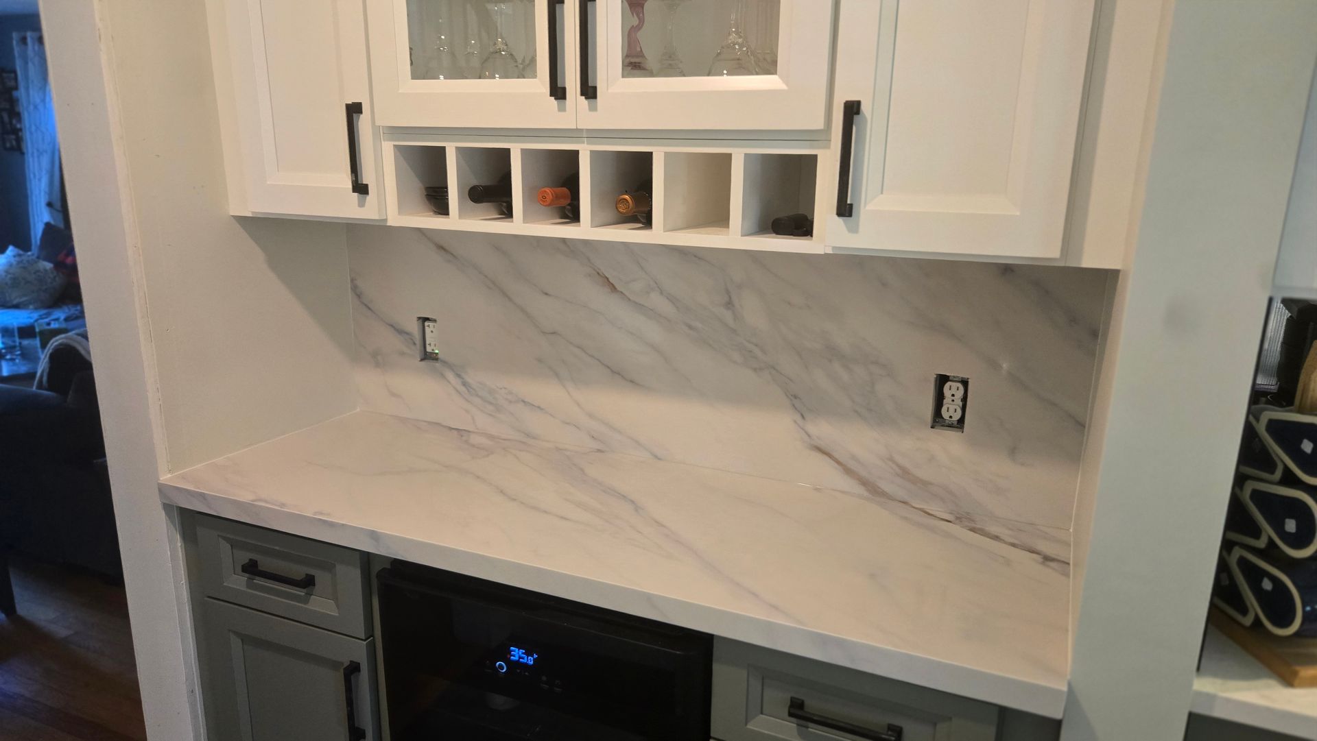 A home wet bar with white cabinetry, a marble-patterned backsplash, and gray lower cabinets surrounding a wine cooler.