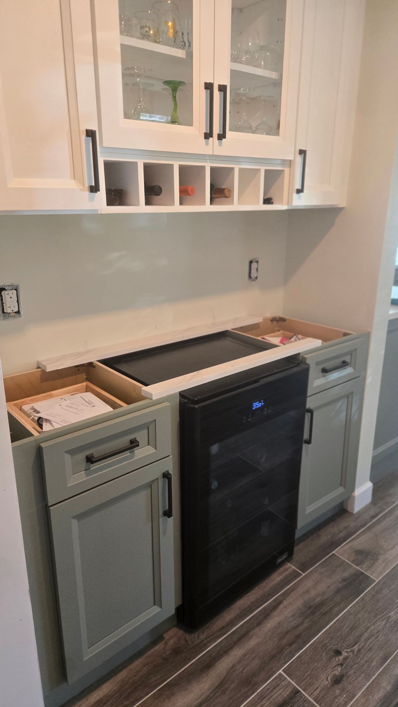 A kitchen bar area with light green base cabinets, a black under-counter beverage cooler, and white upper glass cabinets.