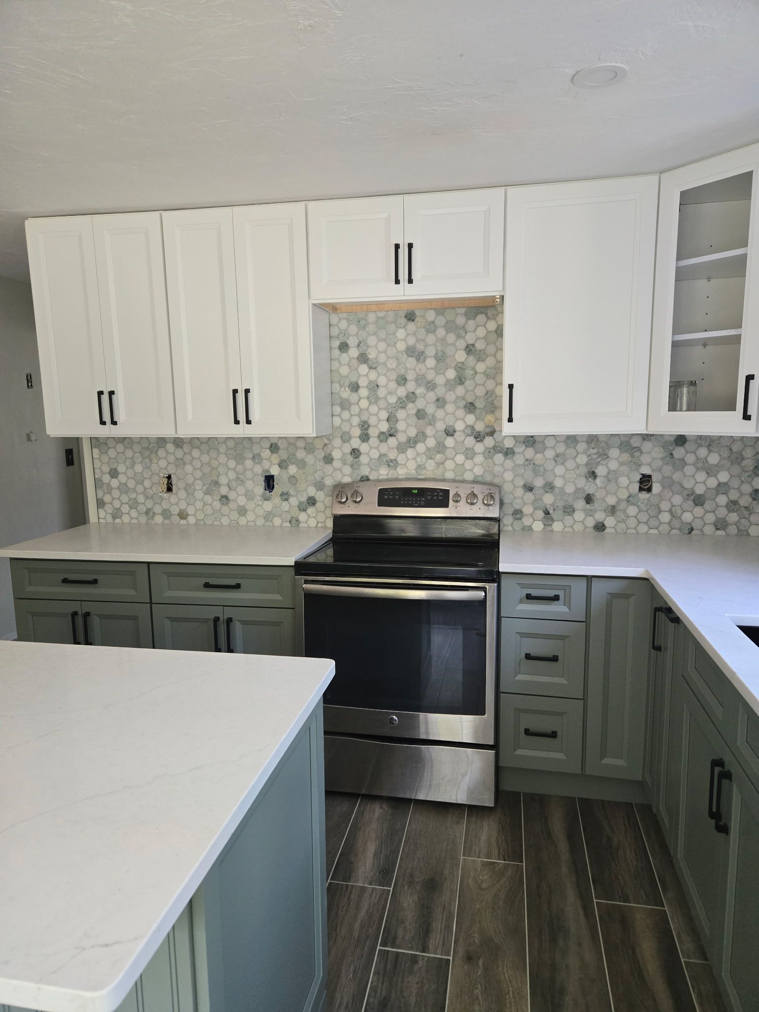 A kitchen with white upper cabinets, green lower cabinets, white countertops, and a patterned mosaic backsplash.