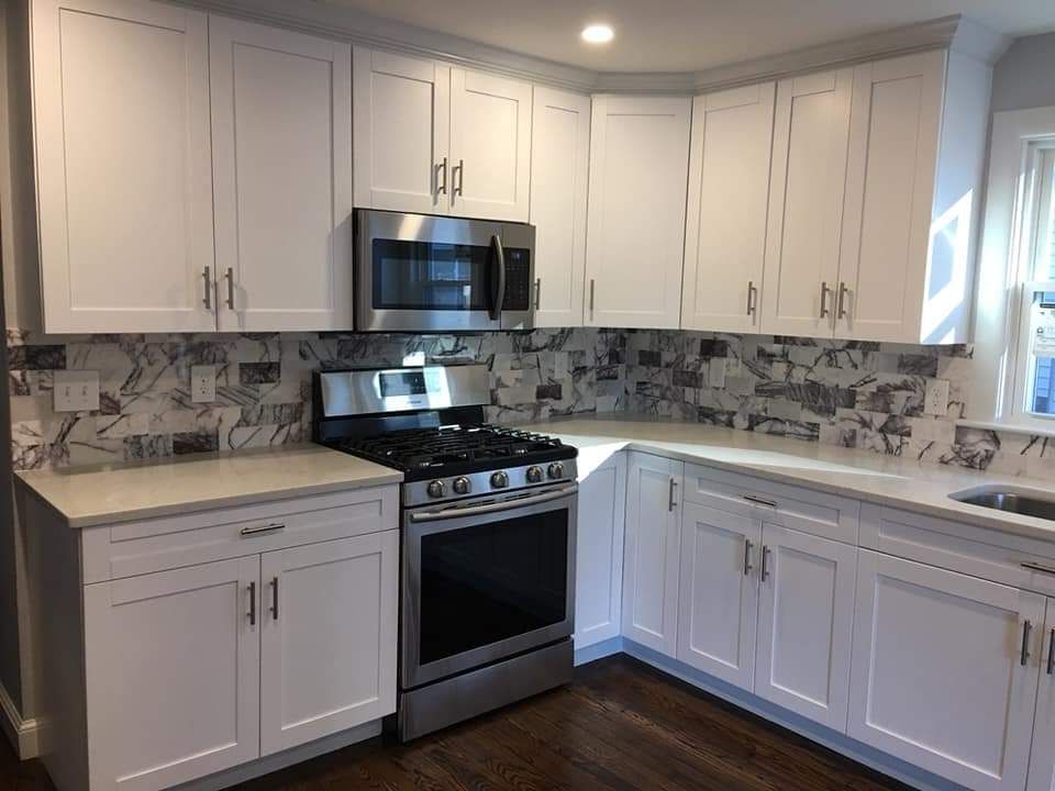 A bright kitchen featuring white cabinets, stainless steel appliances, and a patterned backsplash over dark wood floors.