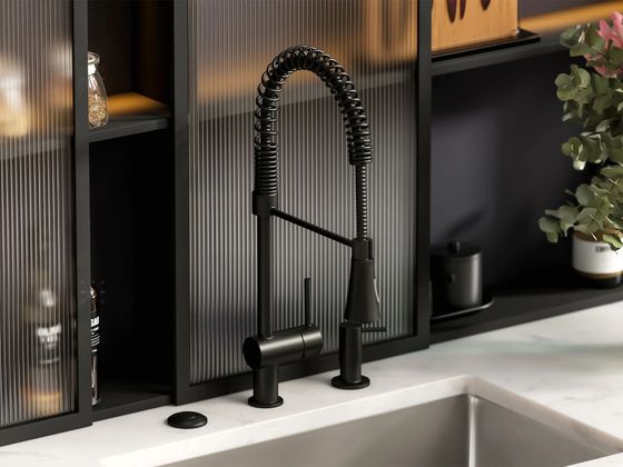 A matte black, spring-neck pull-down kitchen faucet installed on a white countertop in front of fluted glass shelving.