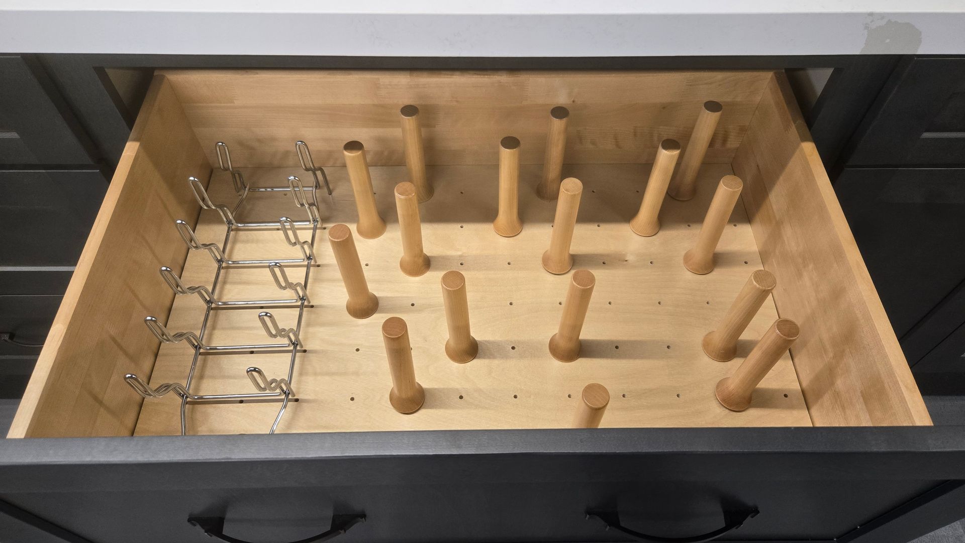 An open kitchen drawer containing a metal plate rack on the left and movable wooden pegs on a grid base for custom storage.