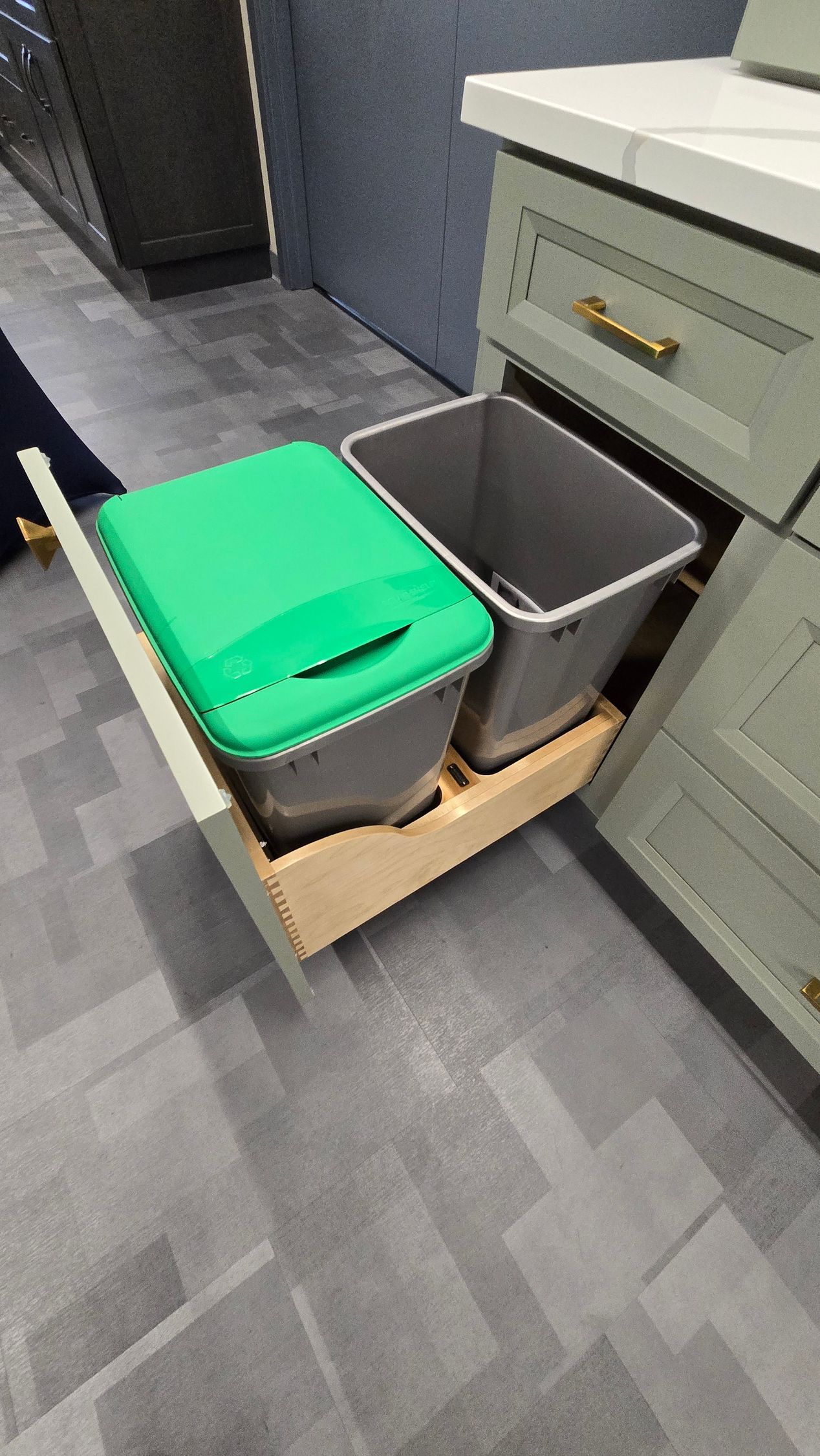 Pull-out kitchen cabinet drawer with two gray trash bins, one with a green lid, inside a light green cabinet base.