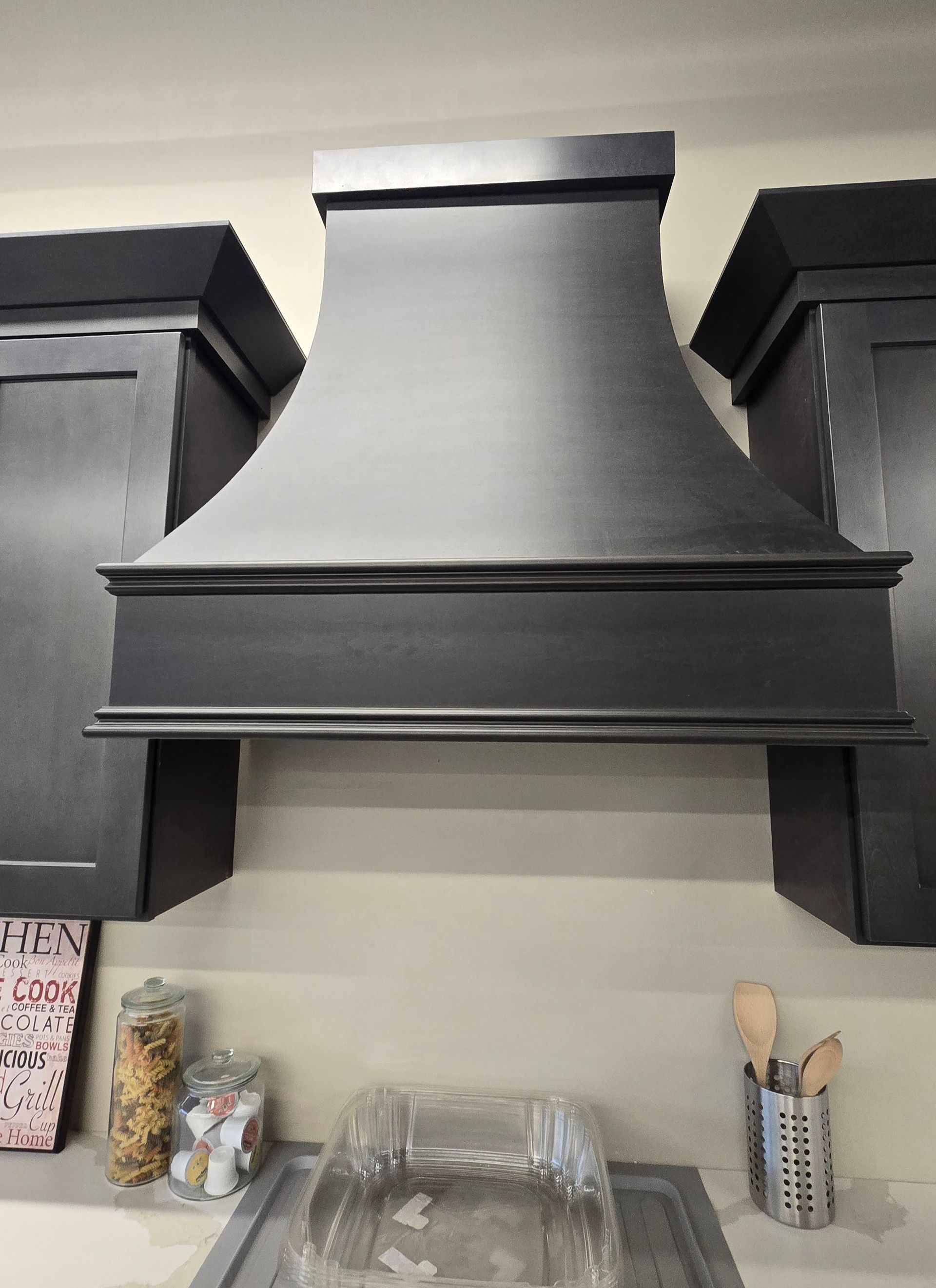 A matte black wooden range hood mounted between two dark cabinets in a kitchen, with kitchen items on the counter below.