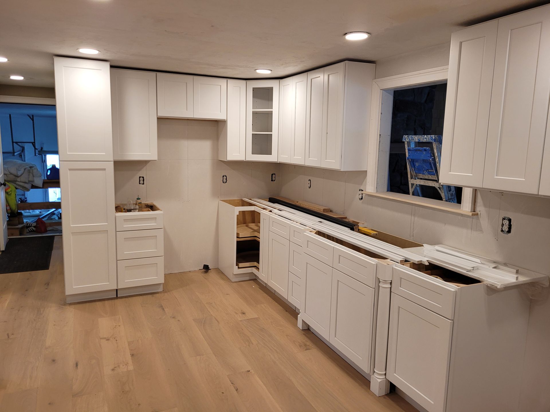 A partially installed white kitchen with light wood floors, base and wall cabinets, and unfinished countertop areas.