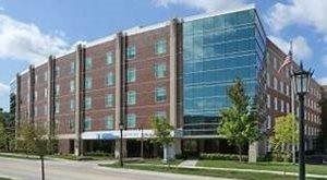 A multi-story brick building with a modern glass facade, featuring an entrance under a blue canopy on a sunny day.