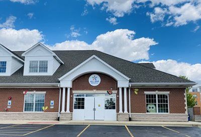A brick building with a white peaked entrance, columns, and a parking lot under a blue sky with scattered clouds.