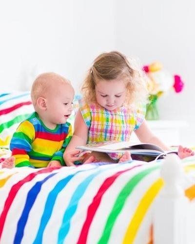 Two children in colorful clothing sit on a rainbow-striped bed reading an open book together.