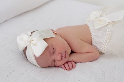 A sleeping baby wears a large cream bow headband and matching ruffled diaper cover with pearl trim on a white blanket.