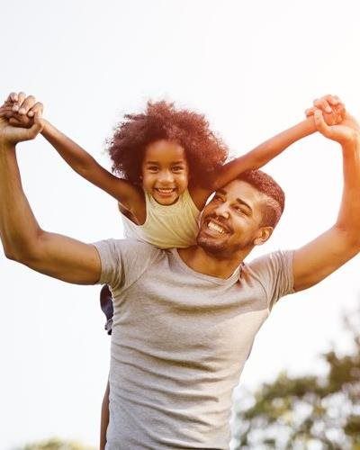 A smiling person gives a child a piggyback ride outdoors, with both reaching their arms up in a celebratory gesture.