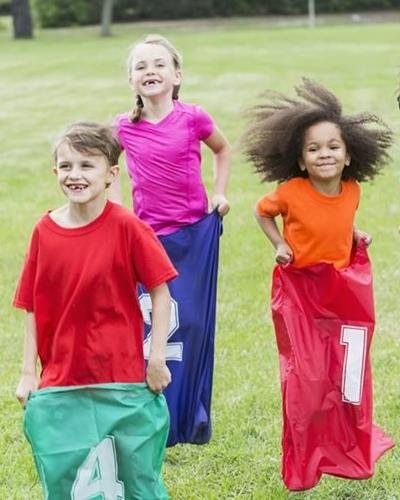 Three children smiling while participating in a sack race on a grassy field, each wearing numbered colorful sacks.