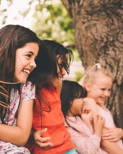 Four happy children laughing while sitting close together outdoors next to a tree.