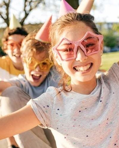 Three smiling people in party hats and fun-shaped glasses pose together outdoors in a sunlit setting.