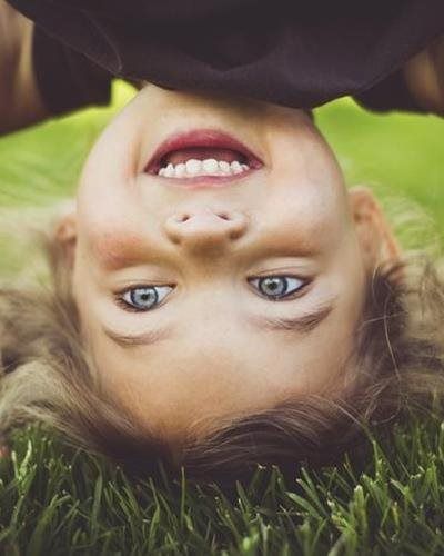A smiling child with bright blue eyes lies upside down in the green grass.