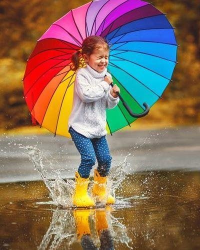 A happy person in a white sweater and yellow rain boots jumps into a puddle, holding a large, colorful rainbow umbrella.