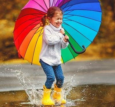 Child in yellow rain boots splashing in puddles under a rainbow umbrella