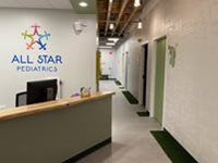Reception desk at All Star Pediatrics with a logo on the wall and a hallway lined with green doors and artificial turf.