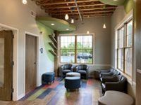 Modern waiting area with blue armchairs and stools, wood-beam ceiling, and large windows with natural light.