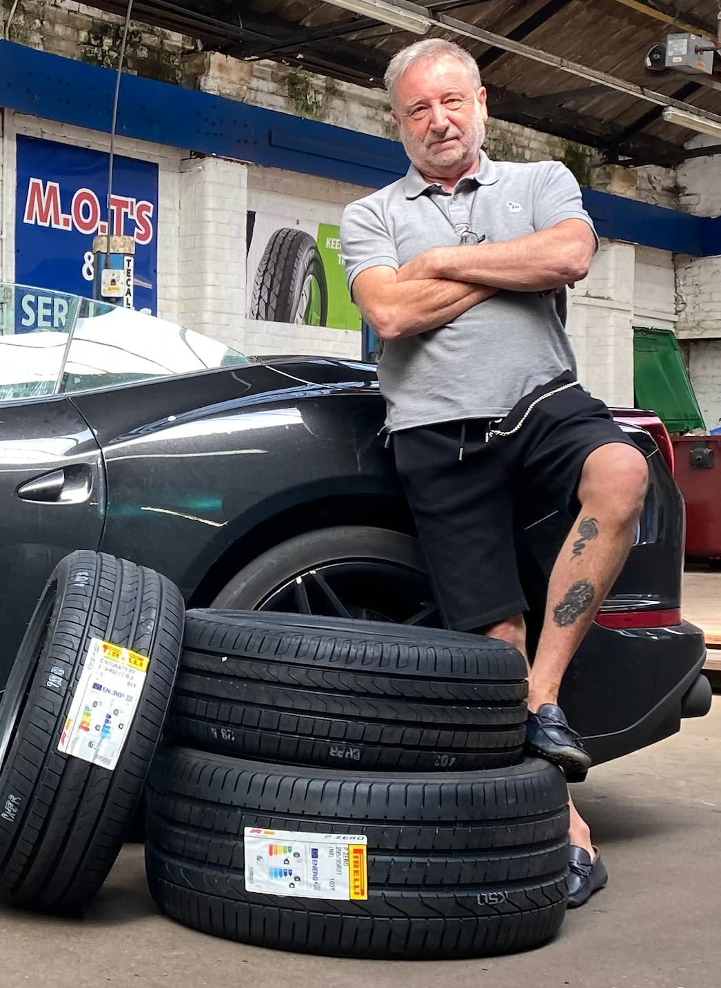 Man with arms crossed leans against car in a garage with tires stacked.