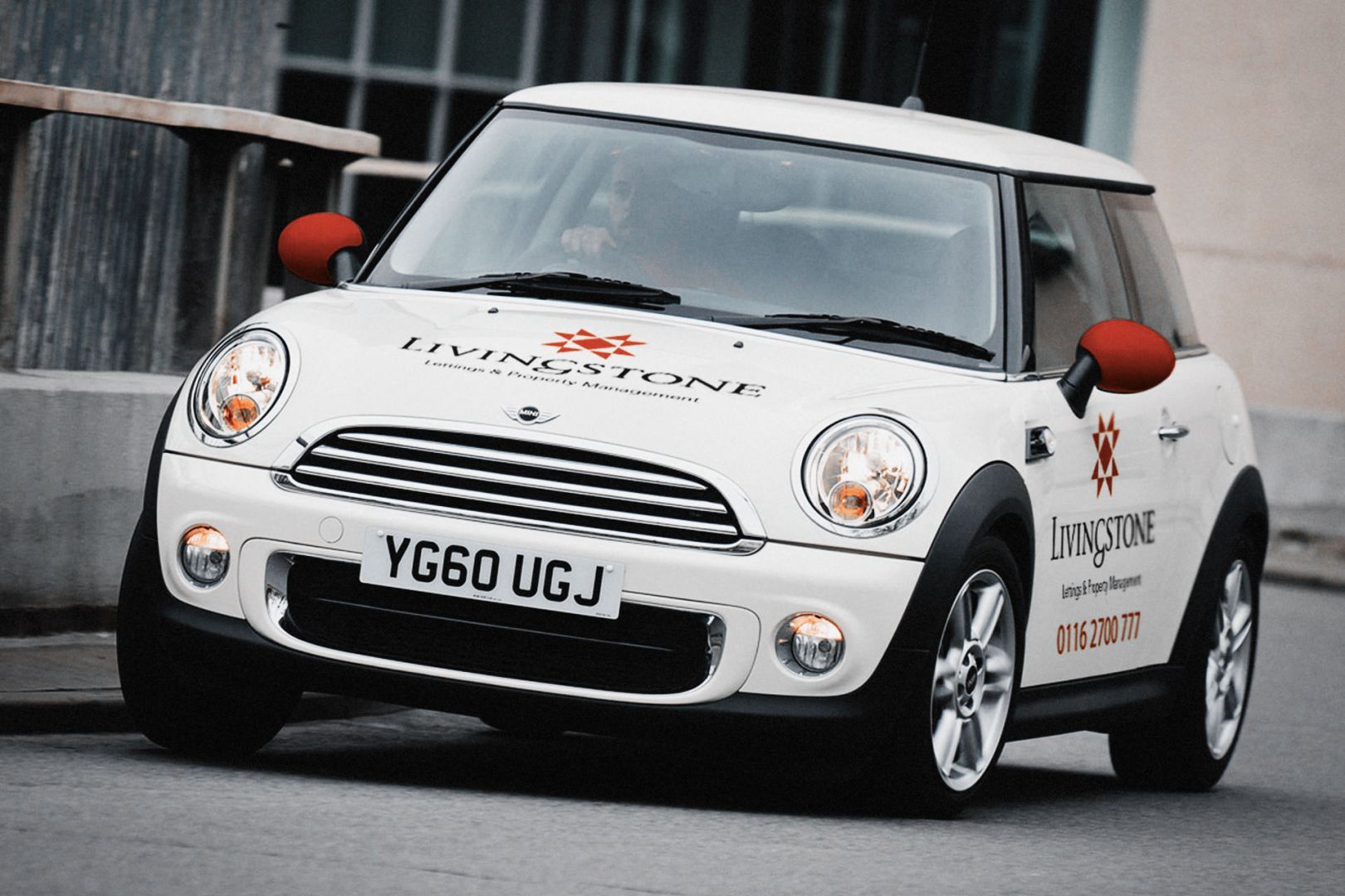 White Mini Cooper with red side mirrors, driving on a road; company logo visible.