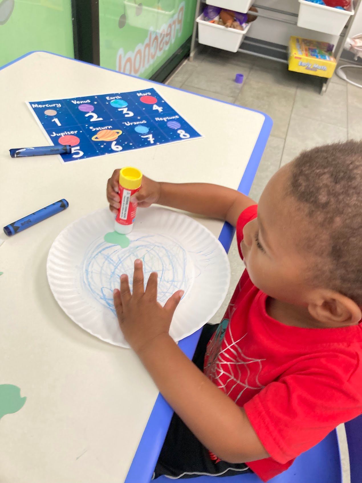 A young boy is sitting at a table using a paper plate to draw a pumpkin