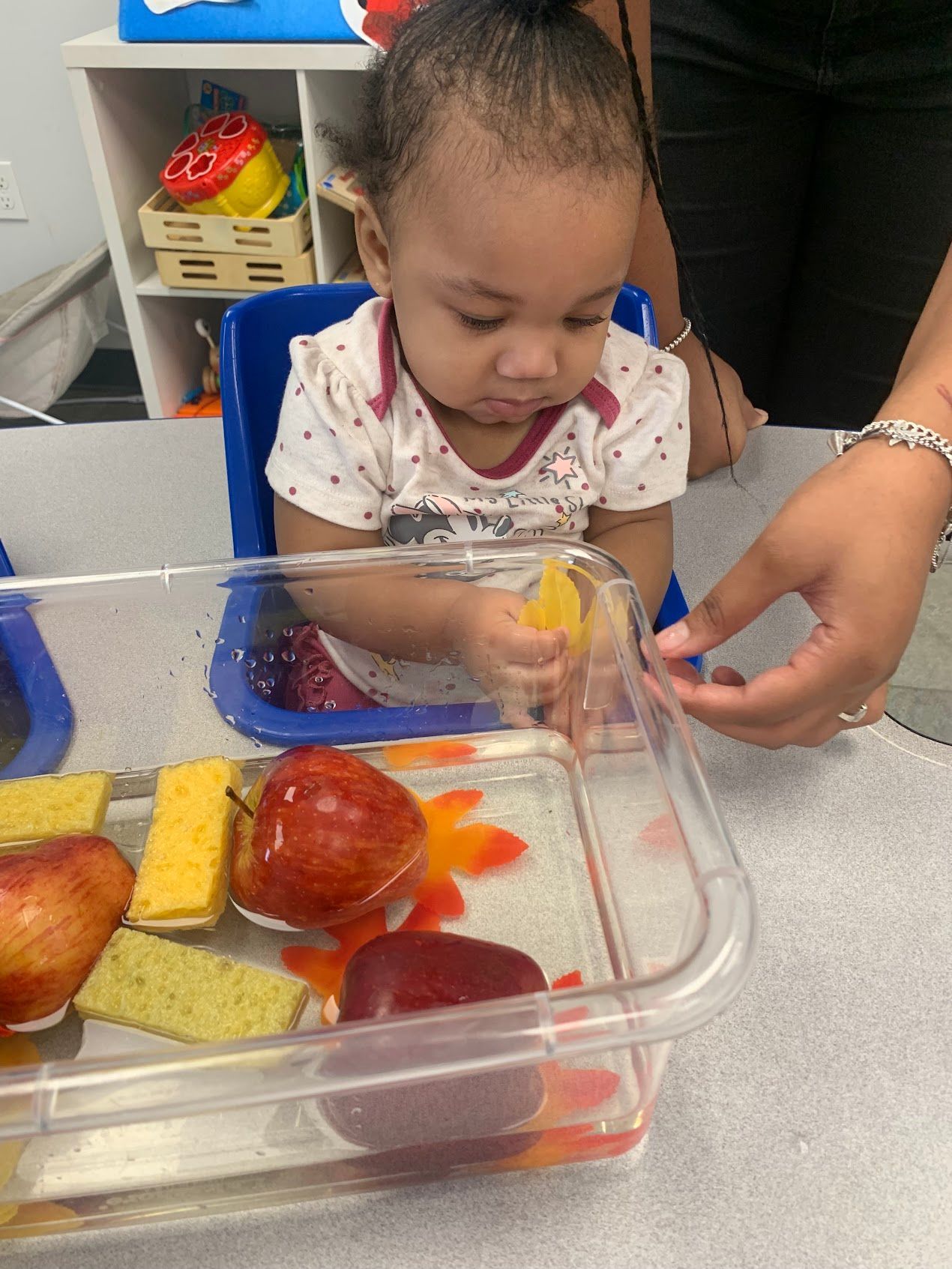 A baby is playing with fruit in a plastic container.