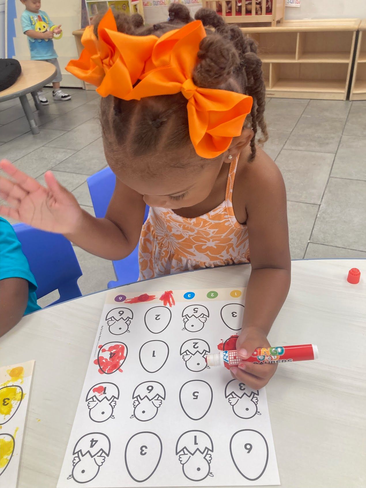 A little girl with an orange bow on her head is drawing on a piece of paper