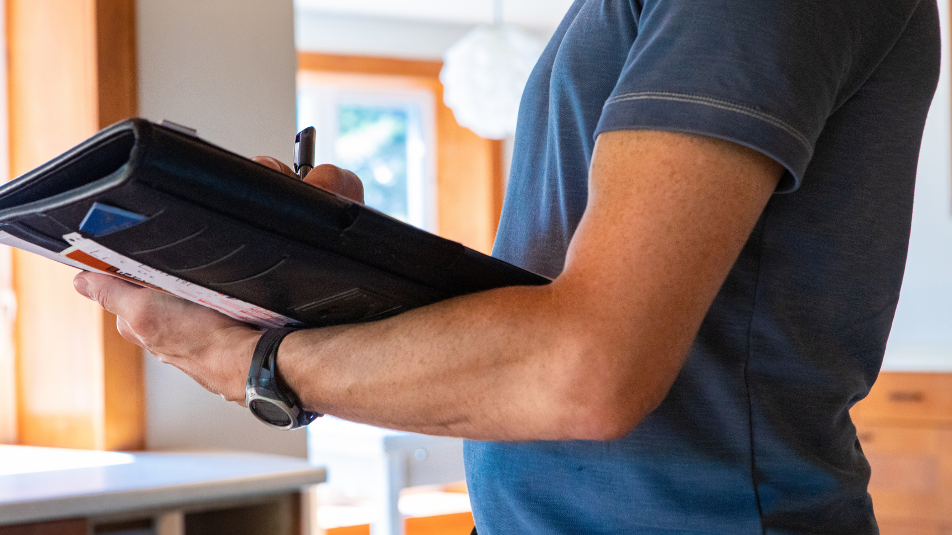 Person in gray shirt holding a binder, writing with a pen. Interior shot with natural light.