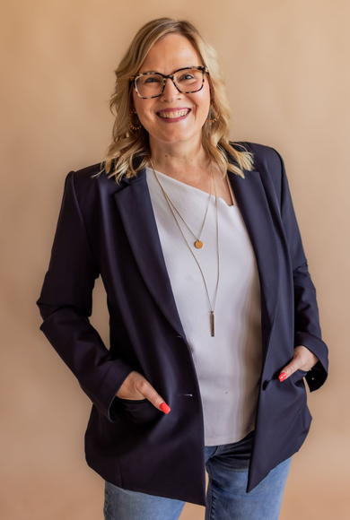 Smiling person with shoulder-length hair and glasses, wearing a dark blue blazer over a white shirt, against a tan backdrop.