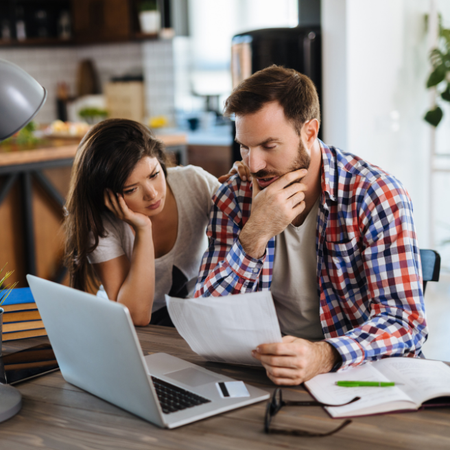 A couple sits at a wooden table looking stressed while reviewing financial documents and a laptop in a home setting.
