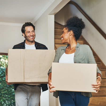 Two people smiling at each other while carrying cardboard boxes into a new home.