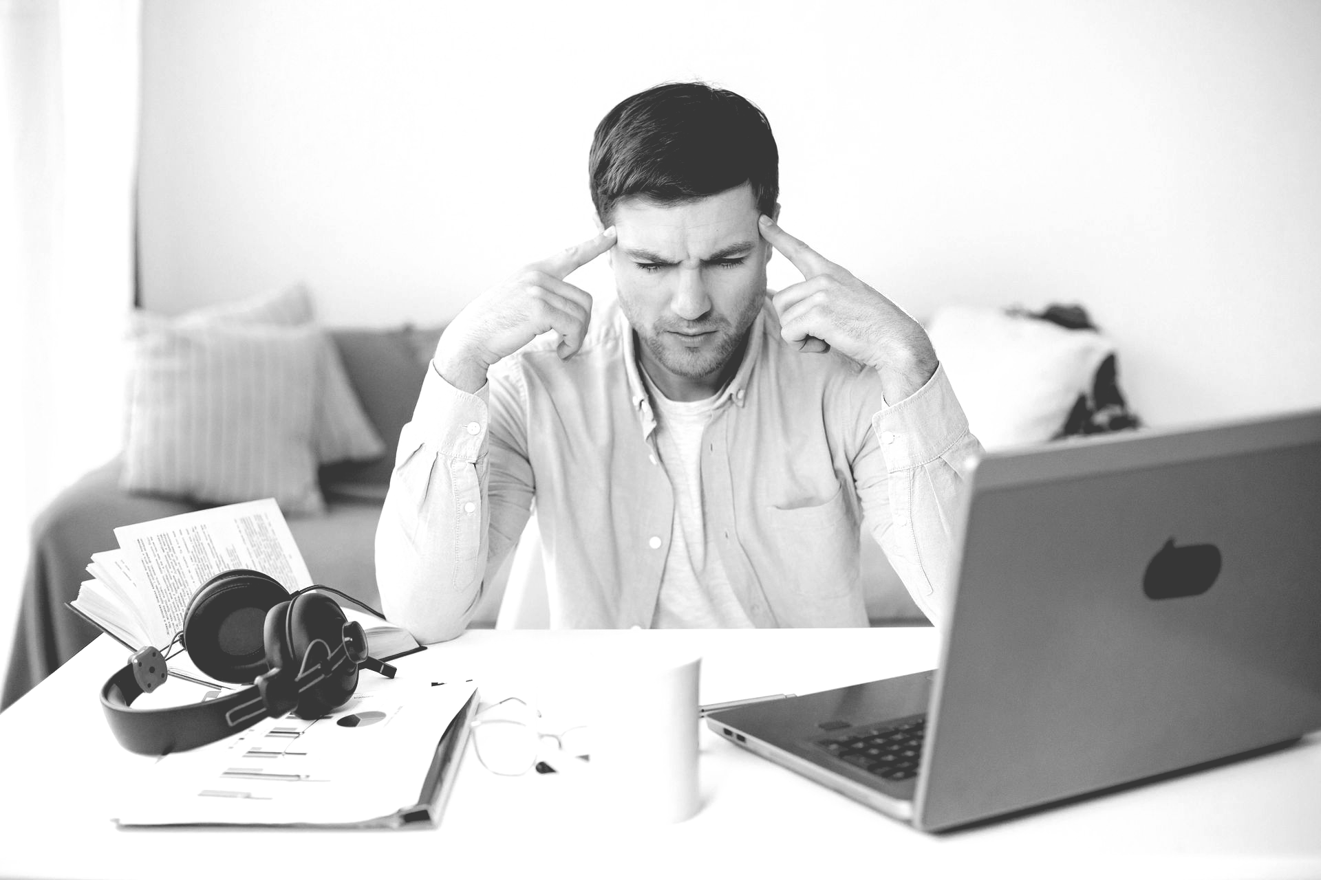 A person sits at a desk with a laptop and headphones, looking stressed with hands held to their temples.