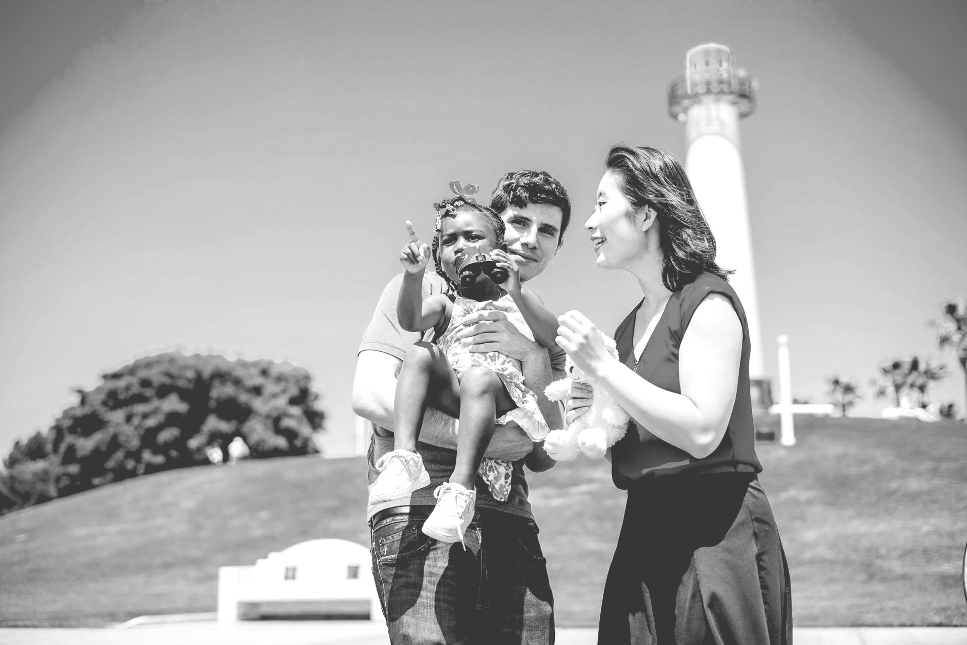 A small child points while being held by a person standing with another individual near a lighthouse.