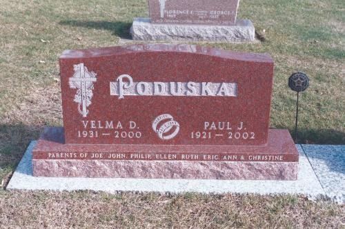 A red granite headstone for Velma D. and Paul J. Paduska, featuring a cross, wedding rings, and their children's names.