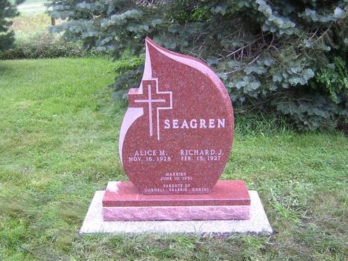 A red granite headstone with a flame-shaped top and a central cross, inscribed with the name SEAGREN and two sets of dates.
