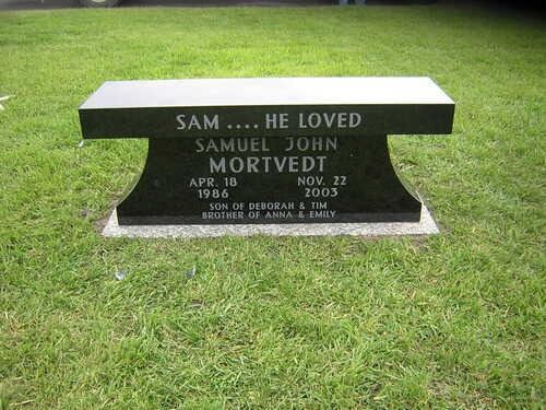 A black granite memorial bench sits on green grass, inscribed with the name Samuel John Mortvedt and his dates of life.