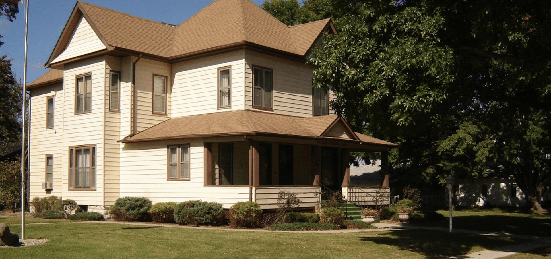 A two-story, cream-colored house with a wrap-around porch and brown roof, surrounded by a green lawn and large trees.