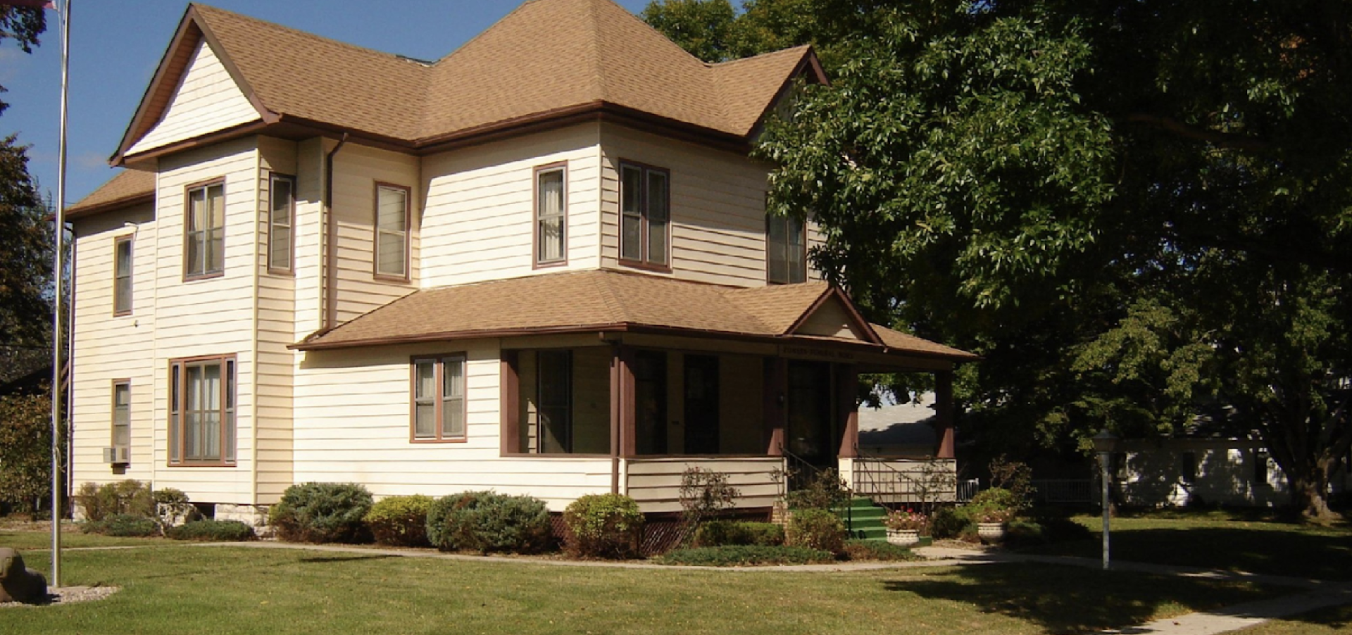 A two-story, cream-colored house with a wrap-around porch and a brown shingled roof, set among trees and green lawns.