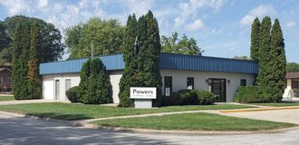 A single-story office building with a blue metal roof, manicured hedges, and a sign reading 