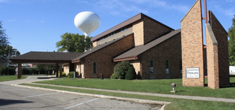A brick building with a brown roof and a tall sign, situated on a grassy lawn with a large white water tower in the back.