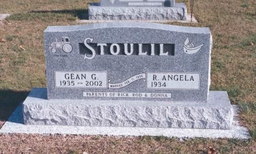 A gray granite headstone for the Stoulil family, featuring engraved tractor and boat icons, set in a grassy cemetery.